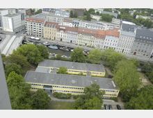 Luftaufnahme einer Stadtansicht: Im Vordergrund ein Gebäudekomplex mit flachen, gelb gestrichenen Gebäuden, umgeben von Bäumen und Grünanlagen. Im Hintergrund eine Reihe mehrstöckiger historischer Fassaden entlang eines Kanals mit festgemachten Booten.