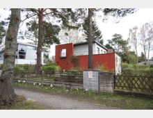 Moderne Wohnhäuser in einer grünen Umgebung, eines mit roten und weißen Fassaden, das andere in hellem Grau. Die Szene zeigt einen gepflasterten Gehweg mit bepflanzten Blumenbeeten im Vordergrund sowie hohe Bäume und eine Mischung aus Holzzäunen und Hecken.