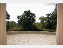 Ansicht eines offenen Parkbereichs mit einer sandigen Lichtung im Vordergrund, umgeben von dichter Vegetation und großen, grünen Bäumen. Zwei weiße Säulen am linken und rechten Bildrand deuten auf einen Rahmen oder einen Portikus hin.