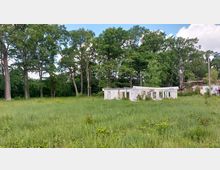 Verlassene Gebäude auf einer überwucherten Wiese, umgeben von hohen Bäumen eines dichten Waldes; die Wände der Gebäude sind mit Graffiti bedeckt und Fensteröffnungen sind leer.