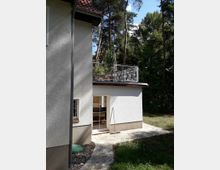 Außenansicht eines Hauses mit hellem Putz und einem roten Ziegeldach, umgeben von hohen Bäumen. Eine Terrasse mit Geländer befindet sich auf einem Flachdachanbau, und eine Glastür führt auf einen kleinen, gepflasterten Bereich im Garten.