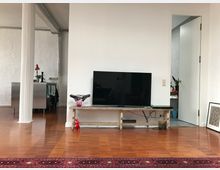 Wohnraum mit glänzendem Holzboden, einer schlichten weißen Wand, an der ein Flachbildfernseher auf einem hölzernen Tisch steht. Links ist eine weiße Säule und ein Bereich mit einer Edelstahlküche sichtbar, rechts führt eine offene Tür in einen weiteren Raum.