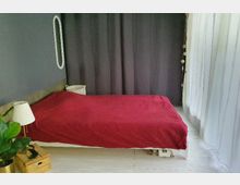 Schlafzimmer mit einem schlichten Bett, das mit einer roten Decke bedeckt ist. Im Hintergrund hängt ein dunkelgrauer Vorhang, rechts sind weiße, lichtdurchlässige Gardinen zu sehen, daneben ein kleiner Nachttisch mit einer Lampe und einer Pflanze.