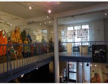 Innenansicht eines Museums mit einer Galerieebene. Links sind Glasvitrinen mit bunten Figuren und Exponaten zu sehen, rechts Fenster und eine Wand mit ausgehängten Fotos. Im unteren Bereich befindet sich ein offener Raum mit weiteren Objekten.