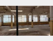 Ehemaliger Industrie- oder Lagerraum mit großen, vergitterten Fenstern, einer hohen Decke mit freiliegenden Rohren und Hängelampen. Die Wände und der Boden zeigen deutliche Abnutzungsspuren, während Säulen den Raum unterteilen.