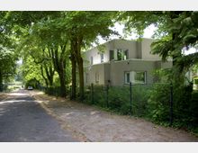 Moderne zweistöckige Wohnhaus mit verputzter Fassade in hellgrauem Ton, umgeben von dichtem Laub und einem schwarzen Metallzaun. Eine schmale Straße führt links am Grundstück entlang, gesäumt von großen Bäumen, im Hintergrund ist ein kleiner See erkennbar.