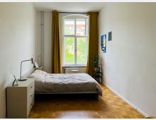 Schlicht eingerichtetes Schlafzimmer mit einem großen Fenster mit gelben Vorhängen, einem Bett mit grauer Bettwäsche, einem Nachttisch mit Lampe und Zimmerpflanze sowie zwei abstrakten Bildern an der Wand. Der Raum hat weiße Wände und einen Parkettboden.