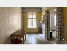 Ein schmaler Raum mit gelben Wänden und floraler Tapete, ausgestattet mit einem Holztisch und zwei Stühlen. Ein großes Fenster mit Blick nach draußen und eine angrenzende, leicht offene Tür mit gemusterten Vorhängen führen in einen weiteren Raum.