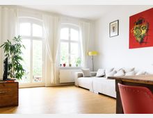 Helles Wohnzimmer mit Holzboden, einer großen beigefarbenen Eckcouch, Gemälden an der weißen Wand und einem roten Stuhl an einem Holztisch. Große Fenster mit weißen Vorhängen lassen Tageslicht herein, und Pflanzen stehen auf der Fensterbank und in der Ecke.