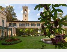 Historischer Innenhof mit gepflegtem Garten, der von einem cremefarbenen Gebäude mit grünen Fensterläden umgeben ist; im Hintergrund ragt ein hoher Turm mit Rundbogenöffnungen empor. Im Vordergrund ranken Passionsblumen, und in der Mitte des Gartens stehen eine Statue und ein kleiner Springbrunnen.