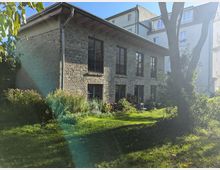 Ziegelsteingebäude mit großen Fenstern und einem abfallenden Holzdach, umgeben von Grünflächen, Sträuchern und Bäumen, im Hintergrund moderne Wohngebäude.