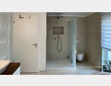 Modernes Badezimmer mit bodengleicher Dusche und Glasabtrennung, wandhängendem WC sowie hellen, neutralen Wand- und Bodenfliesen. Links sind eine weiße Tür und ein Handtuchheizkörper zu sehen, rechts ein großes Fenster mit Rollos und eine Pflanze.