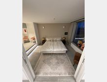 Modernes Schlafzimmer mit einem Doppelbett in der Mitte, flankiert von zwei Nachttischen mit Lampen. Der Raum ist in neutralen Tönen gehalten, umfasst einen großen Fensterbereich mit Vorhängen, einen dekorativen Teppich und einen angrenzenden Bereich mit Sitzmöglichkeiten links.