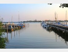 Blick auf einen ruhigen See mit Stegen und kleinen Booten auf beiden Seiten, dahinter eine Küstenlinie mit einer Ortschaft und Kirchtürmen am Horizont. Der Himmel ist hell mit sanftem Abendlicht.
