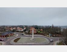 Kreisverkehr in einer Kleinstadt mit einem steinernen Obelisken in der Mitte, umgeben von gepflasterten Straßen und Häusern mit roten Ziegeldächern. Im Hintergrund sind Bäume, eine Kirche und ein See unter einem grauen Himmel zu sehen.
