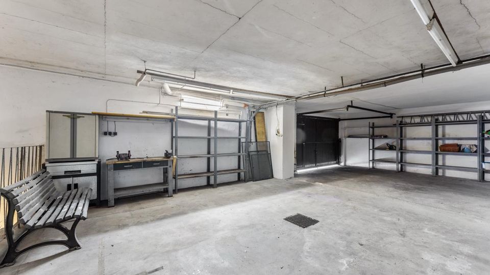 Große, leere Garage oder Lagerraum mit Betonboden und niedriger Decke, ausgestattet mit Metallregalen, Werkbank und Schränken entlang der Wände. An der rechten Seite befindet sich ein breites Tor, in der Decke verlaufen sichtbare Leitungen und Leuchten.