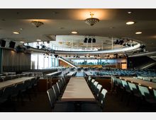 Großer Veranstaltungsraum mit Reihen von langen Tischen und blauen Stühlen, einem erhöhten Rundbalkon mit Geländer, mehreren Deckenlampen und Bühnenscheinwerfern. Im Hintergrund befinden sich große Fenster, eine Treppe und grüne Pflanzen.