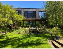 Modernes Wohnhaus mit dunkler Holzfassade und großen Panoramafenstern, umgeben von einem grünen Garten mit Rasen, Bäumen und Pflanzen; eine zentrale Treppe führt vom Garten zur Terrasse des Hauses.