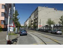 Straßenansicht in einer Stadt mit mehrstöckigen Wohngebäuden auf beiden Seiten, parkenden Autos entlang der Straße und Straßenbahnschienen in der Mitte. Links steht ein Verkehrsschild, und im Hintergrund sind Bäume und weitere Gebäude zu sehen.