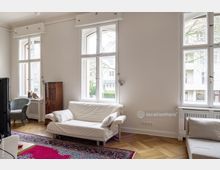 Helles Wohnzimmer mit großen weißen Fenstern, parkettboden, einem weißen Sofa und einem roten Teppich. Im Raum stehen zudem ein geflochtener Stuhl, ein antiker Holzschrank und eine Heizung unter den Fenstern.