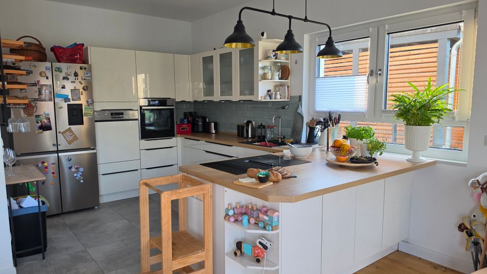 Helle, moderne Wohnküche mit weißen Einbauschränken, Edelstahl-Kühlschrank und einer großen Kücheninsel mit Holzarbeitsplatte und integriertem Kochfeld. Große Fenster mit Blick auf eine Holzverkleidung außen, darüber drei schwarze Pendelleuchten; auf der Insel stehen Schalen, Pflanzen und Küchenutensilien.