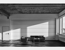 Minimalistisches, modernes Interieur mit einer Wand aus weißen Paneelen, einer schwarzen Ledersitzgruppe mit einem kleinen Tisch sowie einem Sessel. Der Boden ist dunkel, die Decke hat eine sichtbare Betonstruktur, und rechts befindet sich ein großes Fenster, das Tageslicht hereinlässt.