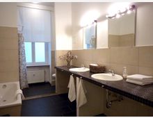 Modernes Badezimmer mit zwei Waschbecken auf einer dunklen gefliesten Arbeitsplatte, darunter Handtuchhalter. Im Hintergrund eine Badewanne mit Duschvorhang und ein Fenster mit halb heruntergelassenem Rollo.