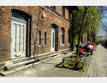 Backsteinfassade eines historischen Gebäudes mit weißen Eingangs- und Fenstertüren, umgeben von einem schmalen gepflasterten Gehweg. Im Vordergrund ein geparkter roter Motorroller und ein rotes Auto neben einem kleinen Baum und einer Sitzbank.