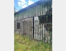 Verwitterter Schuppen aus Wellblech mit einem Holzflügeltor und einem Nummernschild „106“, umgeben von hohem Gras. An der Fassade sind Drahtgitter und teilweise Efeubewuchs zu sehen.