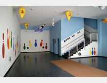 Eingangsbereich eines Bowlingcenters mit bunten Bowling-Pins und Bowlingkugeln als Wanddekoration. Eine Treppe mit Metallgeländer führt nach oben, und an der Decke hängen dekorative Elemente wie Ballons und ein Pfeilschild mit der Aufschrift "Bowling Aufzug".