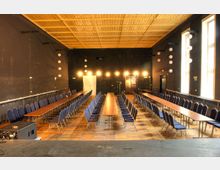 Ein Veranstaltungsraum mit dunklen Wänden, einer Holzdecke und einem Holzboden. Lange Tische sind in Reihen parallel aufgestellt, flankiert von Stuhlreihen, beleuchtet durch kleine Wandlampen und Fenster auf der rechten Seite.