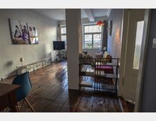 Wohnraum mit Holzboden und heller Wandfarbe, unterteilt durch eine schmale Säule. Im Vordergrund stehen ein Bücherregal und ein kleiner blauer Stuhl, im Hintergrund befinden sich ein Sofa, ein Fernseher auf einem Gestell sowie große Fenster, die Tageslicht hereinlassen.