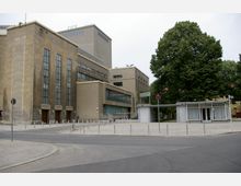 Ein großes, modernes Gebäude im Stil der Nachkriegsmoderne mit hohen rechteckigen Fenstern und einer Fassade aus hellem Stein und Beton. Im Vordergrund ein kleiner Pavillon mit Glasfronten und flachem Dach, daneben ein großer Baum; die Umgebung umfasst gepflasterte Gehwege und eine wenig befahrene Straße.