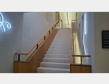 Modernes Treppenhaus in einem öffentlichen Gebäude mit breiter, hellgrauer Steintreppe und Holzgeländern an beiden Seiten. Weiße Wände mit eingelassenen Fensteröffnungen und leuchtenden Wandinstallationen sowie einem Richtungsschild entlang des Treppenaufgangs.