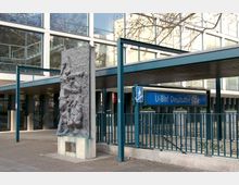 Außenansicht des Eingangs zum U-Bahnhof Deutsche Oper in Berlin, mit einer modernen Glasfassade im Hintergrund und einem Reliefdenkmal im Vordergrund. Ein blaues Schild mit der Aufschrift „U-Bhf Deutsche Oper“ markiert den Zugang zur Treppe.