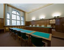 Holzvertäfelter Sitzungssaal mit großen Rundbogenfenstern, einer langen, erhöhten Richterbank und mehreren Tischreihen mit grüner Stoffauflage und Stühlen. Die Wände sind mit Holz verkleidet, und die dekorative Decke ist weiß verziert.