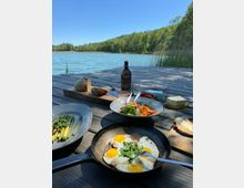 Eine hölzerne Terrasse an einem See, mit Blick auf das Wasser und umgeben von grüner Vegetation. Auf dem Tisch stehen verschiedene Speisen wie Spiegeleier in einer Pfanne, ein Salat, gegrillter Spargel, Brot und Butter, zusammen mit einer Glasflasche und Geschirr.