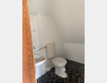 Kleines WC unter einer Dachschräge mit weiß gestrichenen Wänden, Toilette mit Spülkasten und einem kompakten Waschbecken neben einem schmalen Wandschrank. Dunkler Boden aus unregelmäßigen Fliesenfragmenten und Holztürrahmen im Vordergrund.
