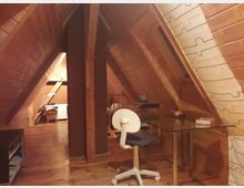 Holzverkleideter Dachbodenraum mit schrägen Wänden, eingerichtet als Arbeitsbereich mit einem Schreibtisch, einem Bürostuhl, einem Regal und einem Fenster. Im Hintergrund befinden sich ein Heizkörper, eine Lampe und ein Stuhl.