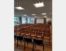 Ein Konferenzraum mit mehreren Reihen aus Holzstühlen, ergänzt durch einige schwarze Kunststoffstühle im Vordergrund. Im Hintergrund befinden sich eine Leinwand, Vorhänge und große Fenster, durch die Tageslicht einfällt.