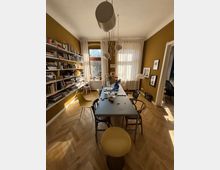 Sunlit dining room with mustard-colored walls, a herringbone wood floor, and a long rectangular table surrounded by mismatched chairs beneath modern pendant lights. A wall-length bookshelf lines one side, and large windows with sheer curtains face the table, with framed artwork and doorways to adjacent rooms on the right.