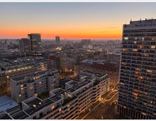 Stadtansicht bei Sonnenuntergang mit mehreren modernen und älteren Gebäuden, teilweise beleuchtet, sowie einem hohen Wohnhochhaus im Vordergrund rechts. Der Himmel zeigt einen orangefarbenen Streifen über der abklingenden Dämmerung.