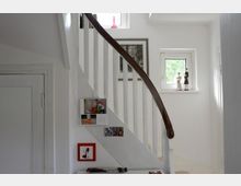 Treppenhaus in einem privaten Wohnhaus mit einer weißen Holztreppe und einem dunklen, geschwungenen Handlauf. An der Wand befinden sich Bilder und Dekorationen, während kleine Fenster natürliches Licht hereinlassen.