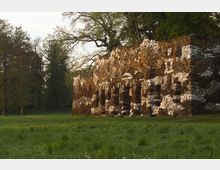 Ruine einer historischen Steinstruktur inmitten einer grünen Wiese, umgeben von hohen Bäumen. Die Fassade besteht aus Bruchsteinen in verschiedenen Farbtönen und zeigt mehrere bogenförmige Öffnungen.