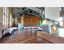 Innenansicht eines Veranstaltungsraums mit Backsteinwänden, Holzboden und einer offenen Decke mit sichtbaren Stahlträgern. Im Raum stehen runde Stehtische mit roten Tischdecken sowie ein holzverkleidetes Rednerpult neben einer großen, dekorierten Pflanze.