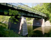 Eine grüne Stahlbrücke mit dekorativen Geländern überspannt einen Fluss, gestützt von massiven Pfeilern aus roten Ziegeln und Beton. Im Hintergrund ist dichter Baumbestand erkennbar, während im Vordergrund Pflanzen heranwachsen.