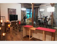 Ein Studio-Set mit Holzmöbeln, hellem Holzboden und sichtbaren Backsteinwänden. Im Raum befinden sich Kameras auf Stativen, Studioleuchten, ein Bücherregal mit Deko und ein Fernseher; im Hintergrund ist ein großes Fenster sichtbar.