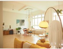 Heller Wohnraum mit modernem, offenem Design, gelbem Sofa, geschwungener Stehleuchte, Esstisch mit Stühlen und großen Fenstern, die viel Tageslicht einlassen. An der Wand befindet sich eine Kommode und Dekoration, darunter Pflanzen und gerahmte Bilder.