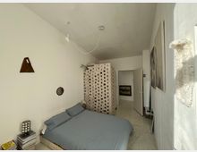 Ein schlicht eingerichtetes Schlafzimmer mit einem Bett mit hellblauer Bettwäsche, einer gemauerten, durchbrochenen Raumtrennung und weißen Wänden. Im Raum befinden sich minimalistische Dekorationen wie ein kleiner runder Spiegel, eine Wandlampe und eine aufeinander gestapelte Sammlung von Büchern oder Magazinen.