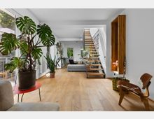 Wohnzimmer in modernem Design mit Holzboden, großen Fenstern und vielen Pflanzen. Eine freitragende Holztreppe mit Metallgeländer führt zu einem Bücherregal, während zahlreiche Möbel und Dekorationen den offenen Raum ergänzen.